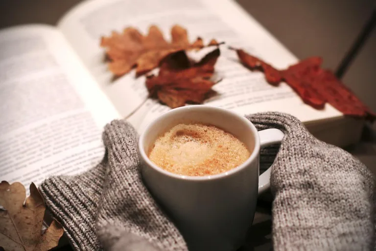 A cup of coffee next to a book with autumn leaves, representing autumn self-care ideas.